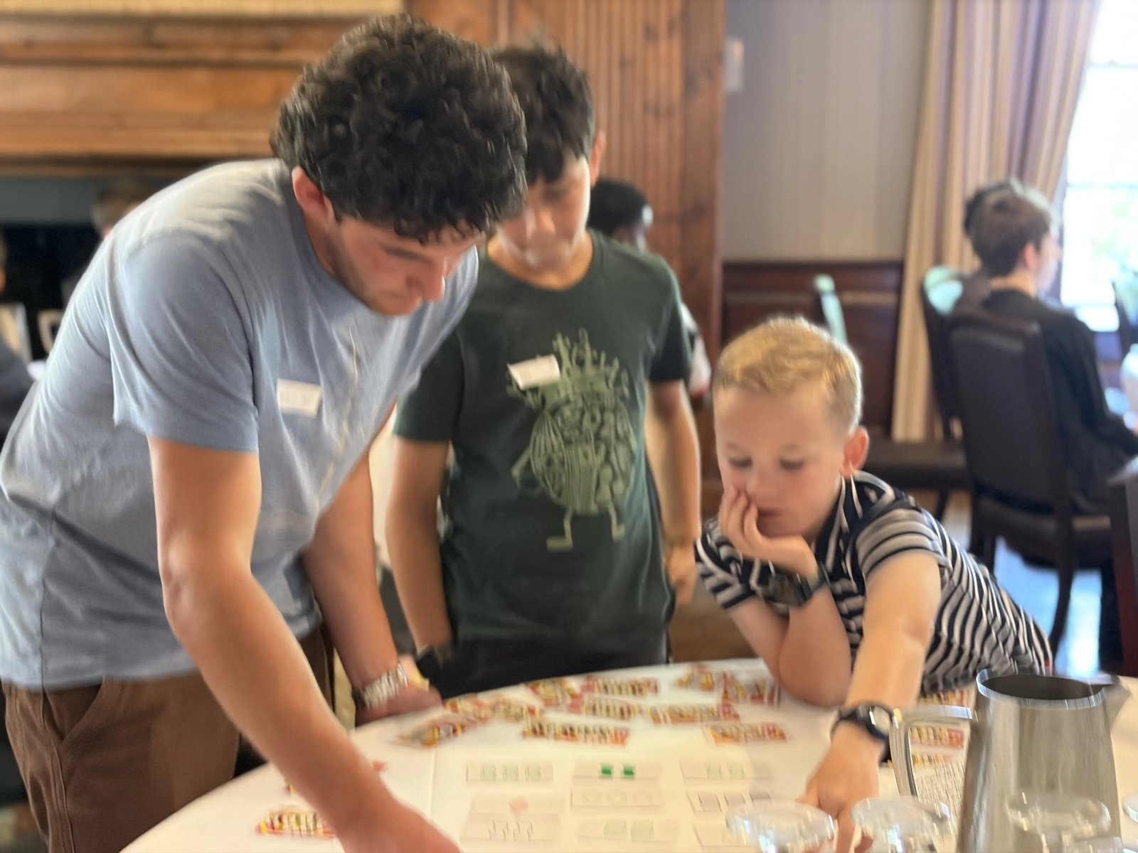 A CyberMath instructor leans over a table with two younger students as they work through a card-based logic puzzle