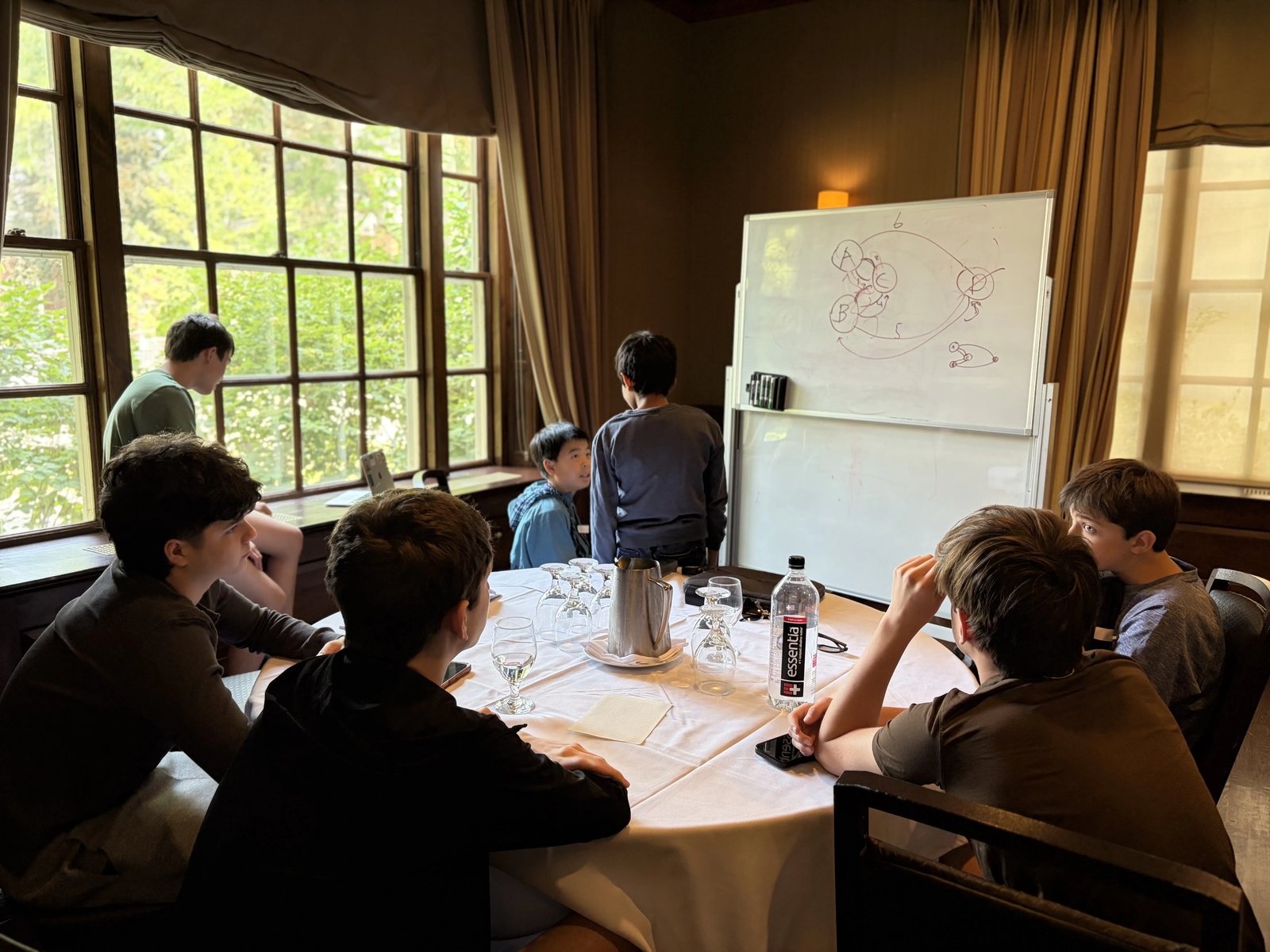 Students talk through a diagram on a whiteboard around a table in a wood-panelled room with tall windows