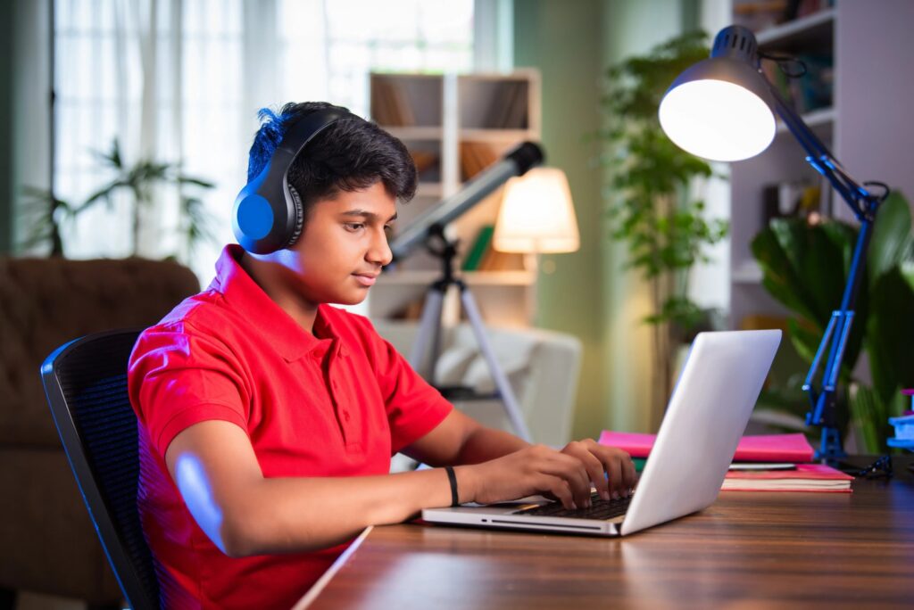 A teenage student wearing headphones works on a laptop at a desk at home during an online lesson